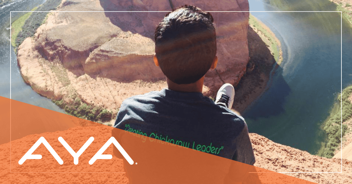 Person wearing 'Creating Chickasaw Leaders' t-shirt sits overlooking a dramatic red rock canyon with blue water below. View is from behind, showing AYA logo with coral overlay. Image illustrates connection between nature walks and mindfulness.