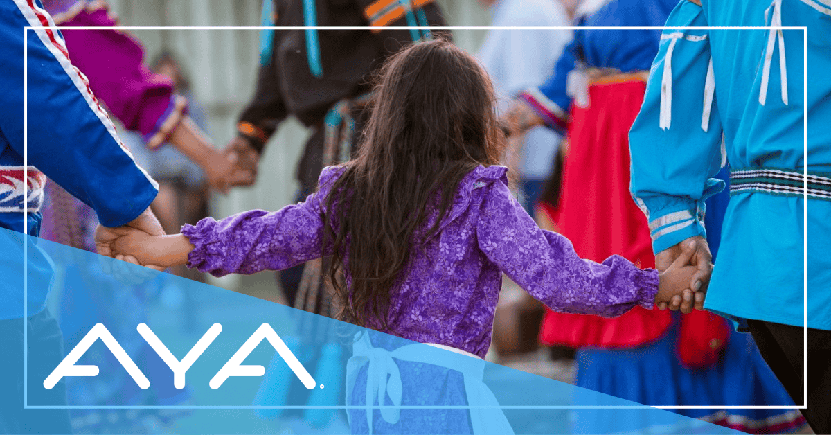Dancers hold hands during a traditional Chickasaw dance, with a young girl in a purple ribbon dress seen from behind, holding hands with others wearing blue ribbon shirts. Image includes a blue diagonal overlay with 'AYA' logo in white.