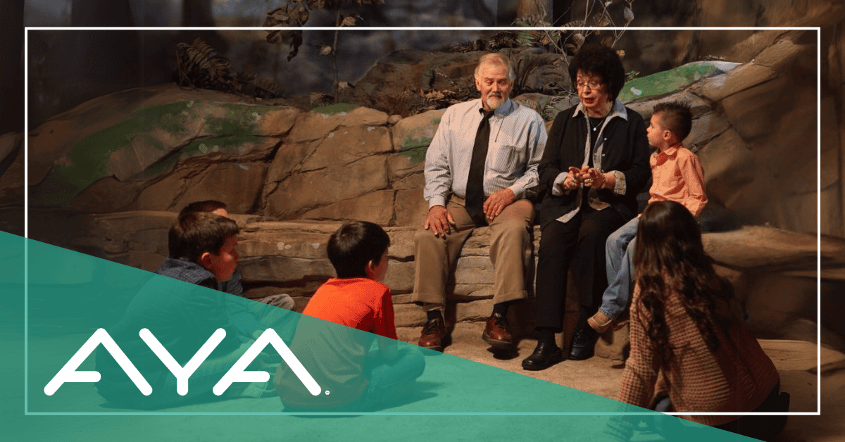 A grandmother sits with a child beside her, telling stories as four other children listen attentively around her. The atmosphere is warm and inviting, with the children fully engaged in her storytelling, creating a sense of connection and tradition.